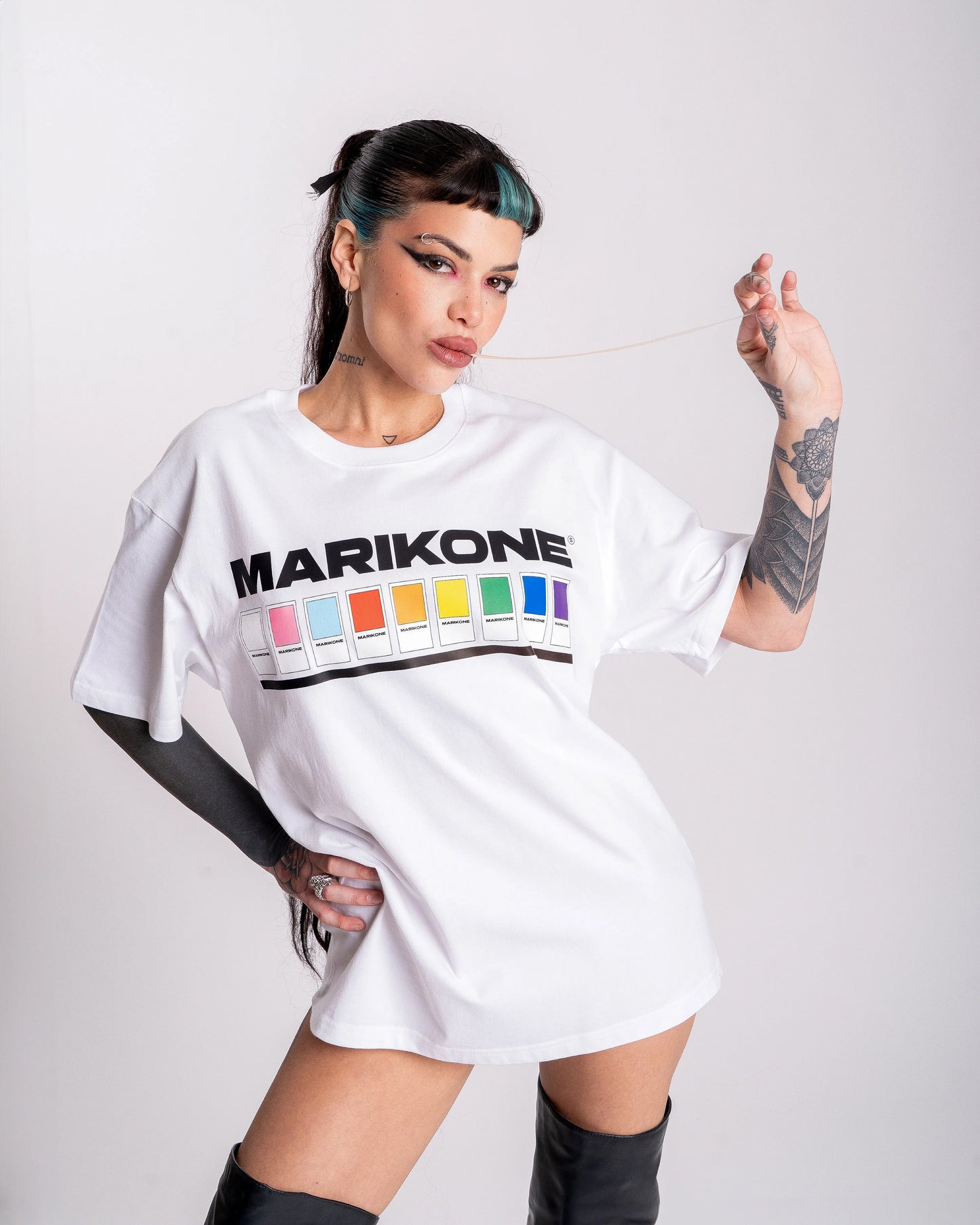 Chica con coleta larga estirando un chicle con su mano, posando con una camiseta con colores de bandera gay lgtb, trans y queer con fichas de color como pantone y que dice MARIKONE