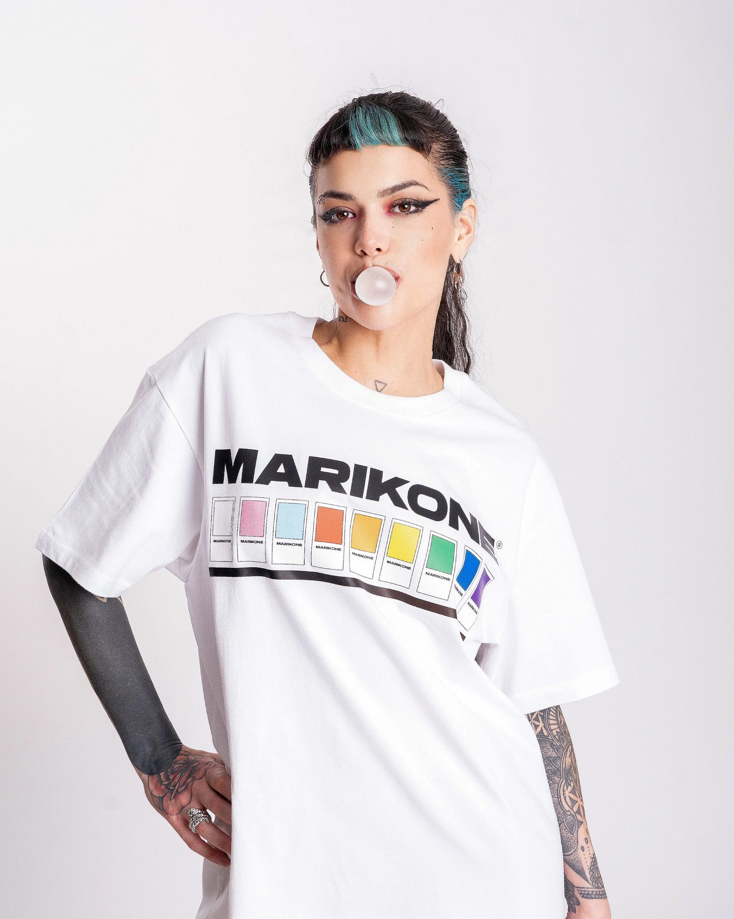 Chica haciendo pompa de chicle. Lleva una camiseta con colores de bandera gay lgtb,queer y trans con fichas de color como pantone que dice MARIKONE