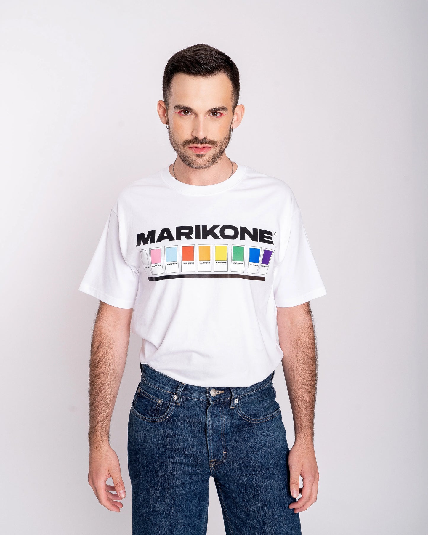 Chico con sombra de ojos rosa posando de frente con una camiseta con colores de bandera gay lgtb, trans y queer con fichas de color como pantone y que dice MARIKONE