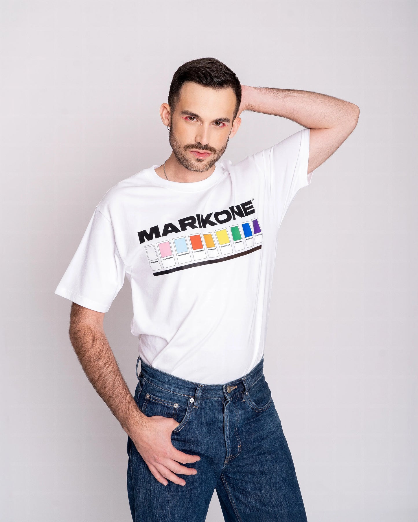 Chico con sombra de ojos rosa posando de frente con la con la mano detrás de la cabeza y llevando una camiseta con colores de bandera gay lgtb, trans y queer con fichas de color como pantone y que dice MARIKONE