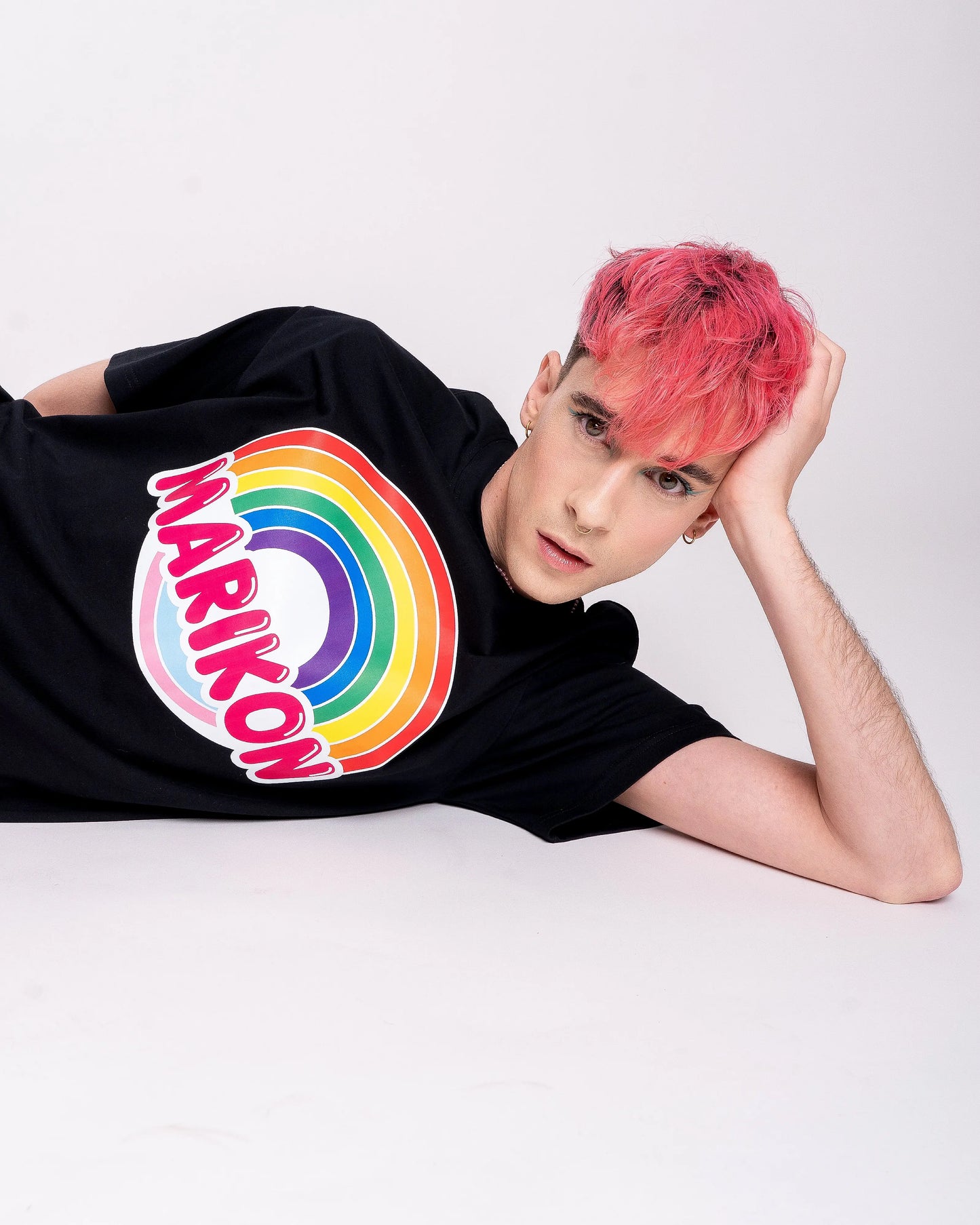 Un chico con cabello rosa recostado en el suelo con la mano sobre la cabeza. Lleva camiseta negra con motivo MARIKON, con arcoíris y colores de bandera trans. Camiseta del orgullo LGTBIQ+ y Queer