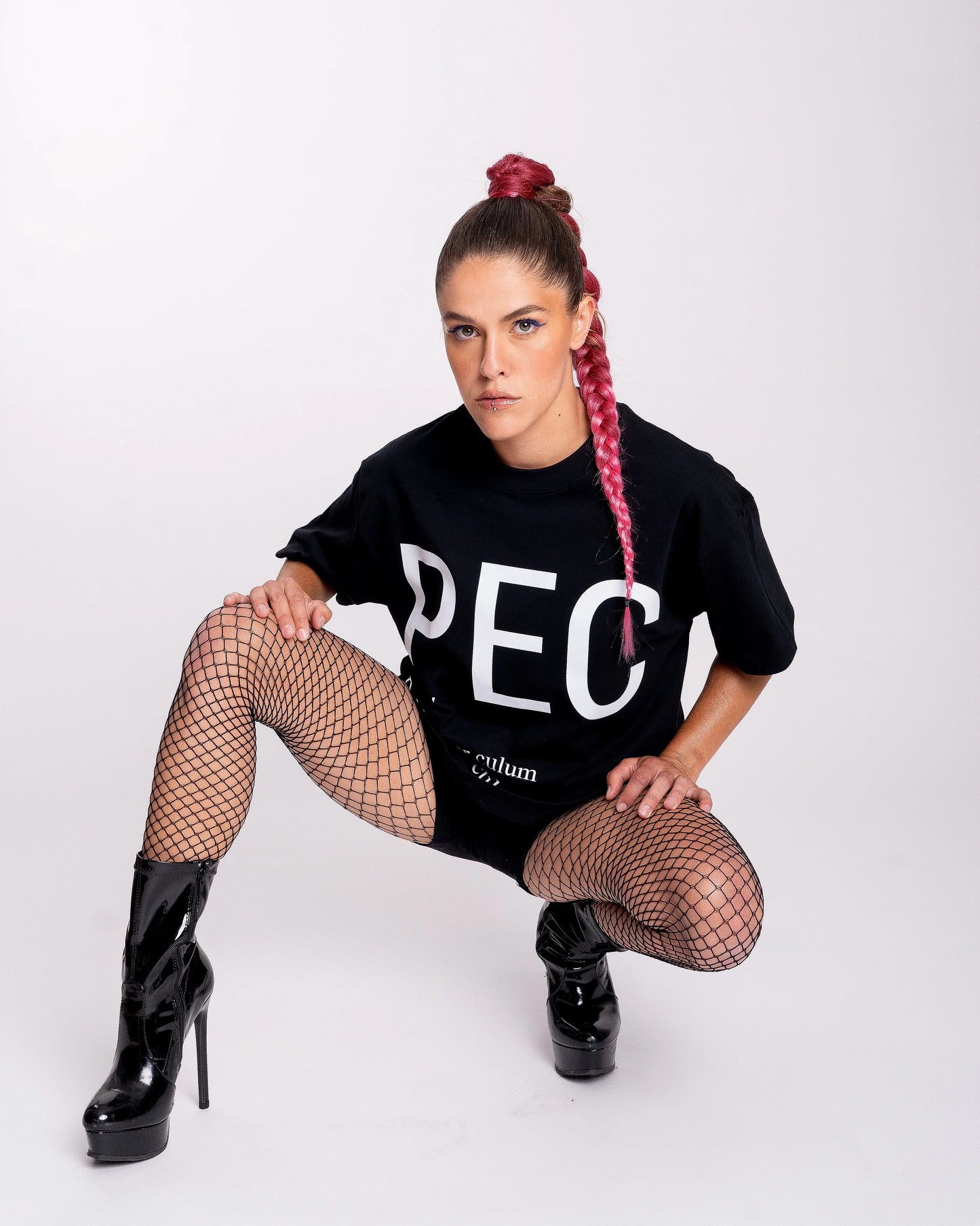chica queer agachada con botas de tacón negro y mallas posando con camiseta negra con letras blancas grandes que dicen PEC, definición: por el culo. Una expresión del colectivo LGTBIQ+ / gay
