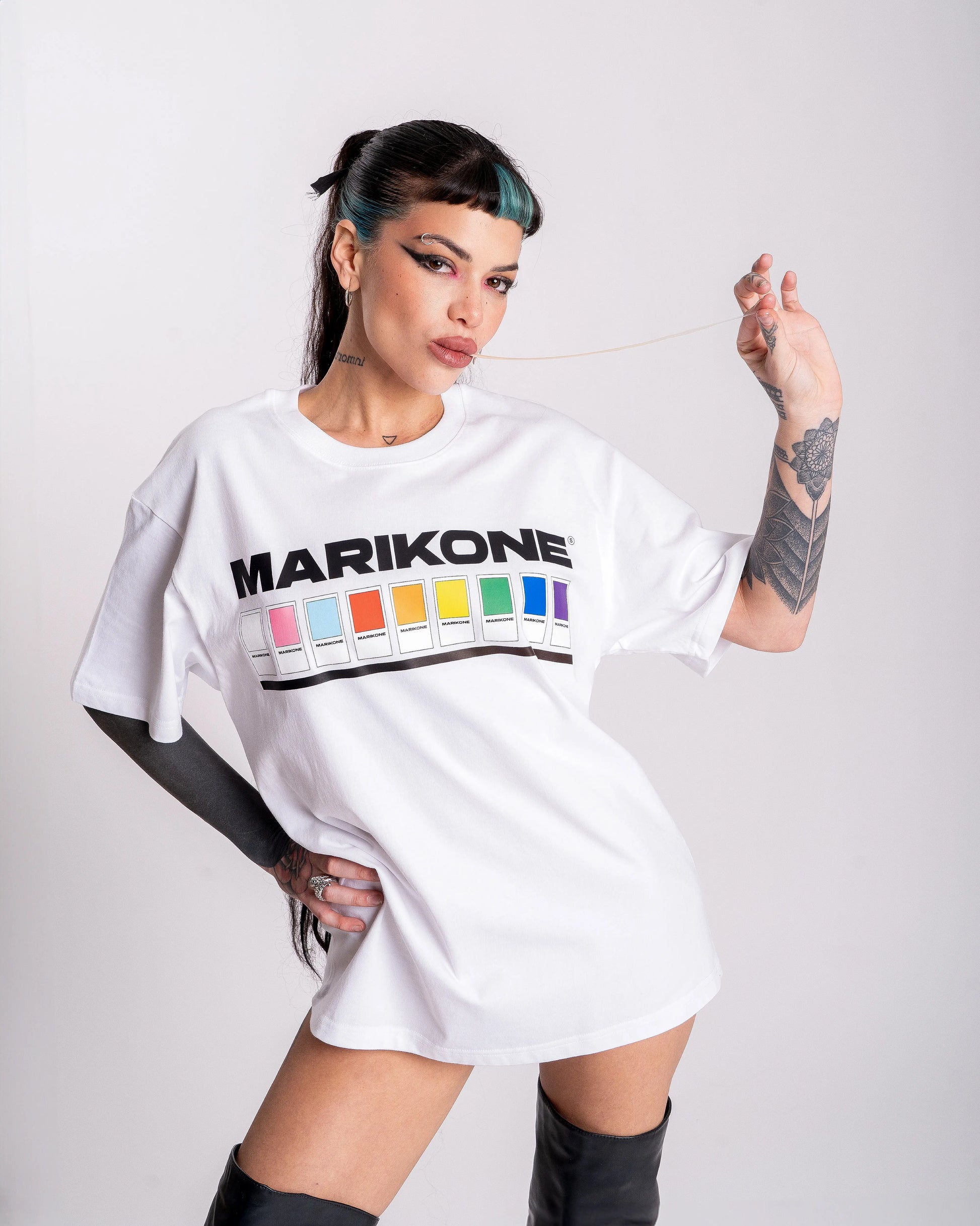 Chica con coleta larga estirando un chicle con su mano, posando con una camiseta con colores de bandera gay lgtb, trans y queer con fichas de color como pantone y que dice MARIKONE