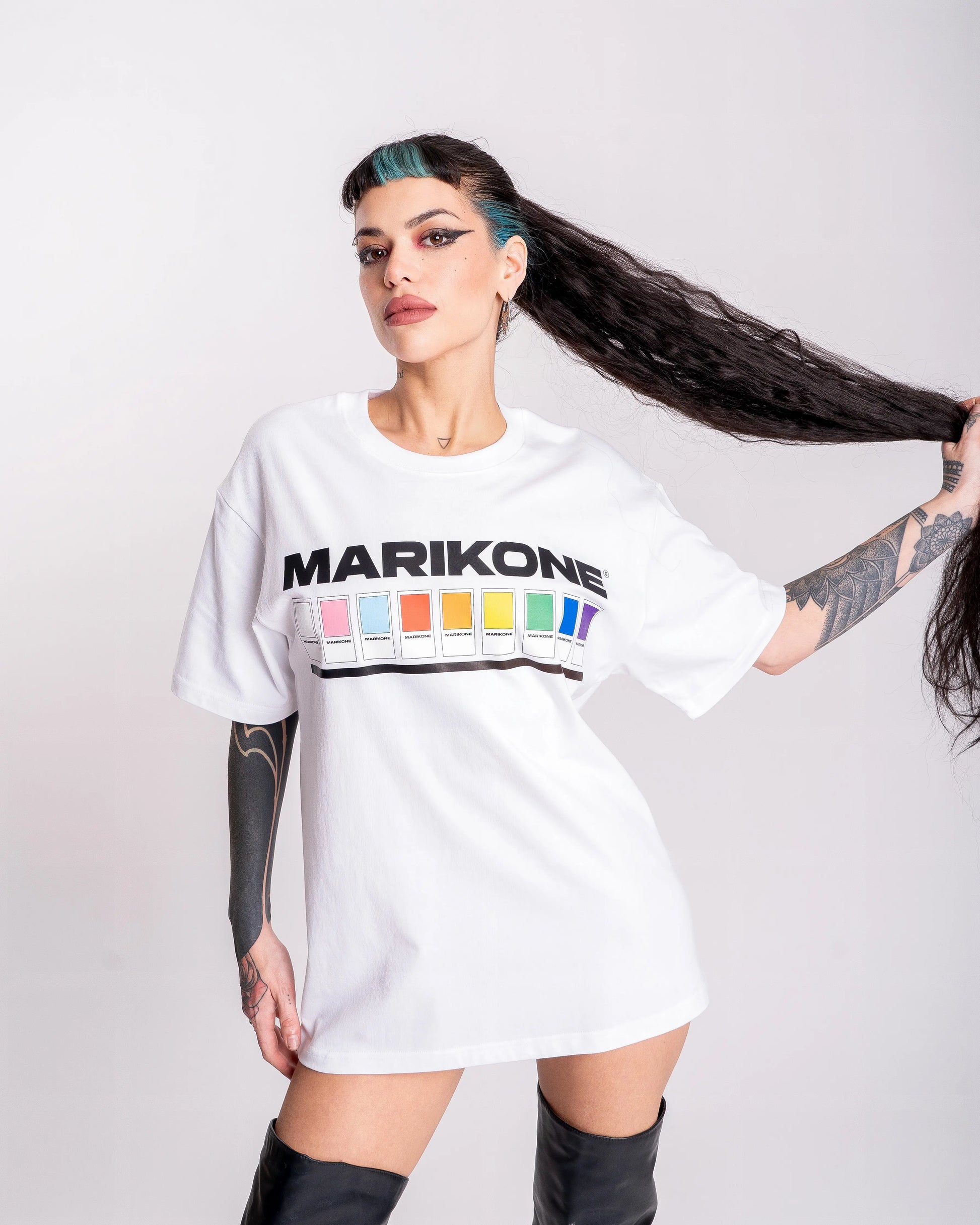 Chica con coleta larga cogiéndola con una mano, pasa con camiseta con bandera gay lgtb, trans y queer con catalogo de colores como pantone y que dice a su vez  MARIKONE.