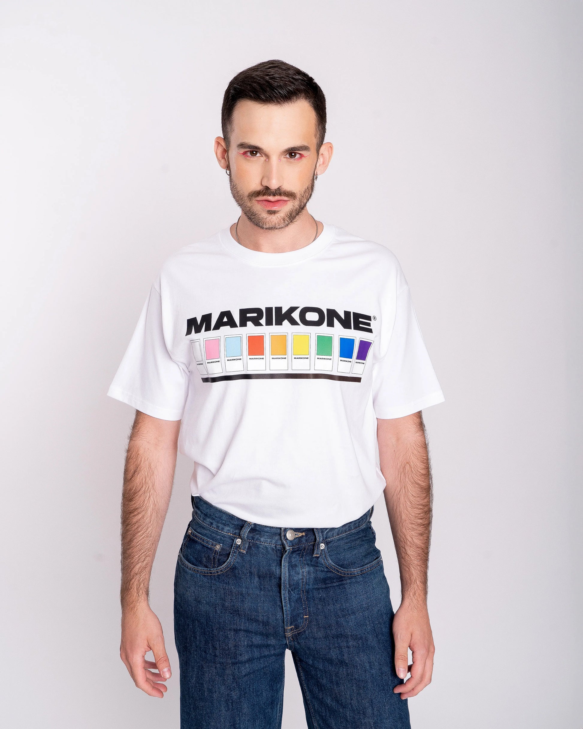 Chico con sombra de ojos rosa  posando de frente con una camiseta con colores de bandera gay lgtb, trans y queer con fichas de color como pantone y que dice MARIKONE