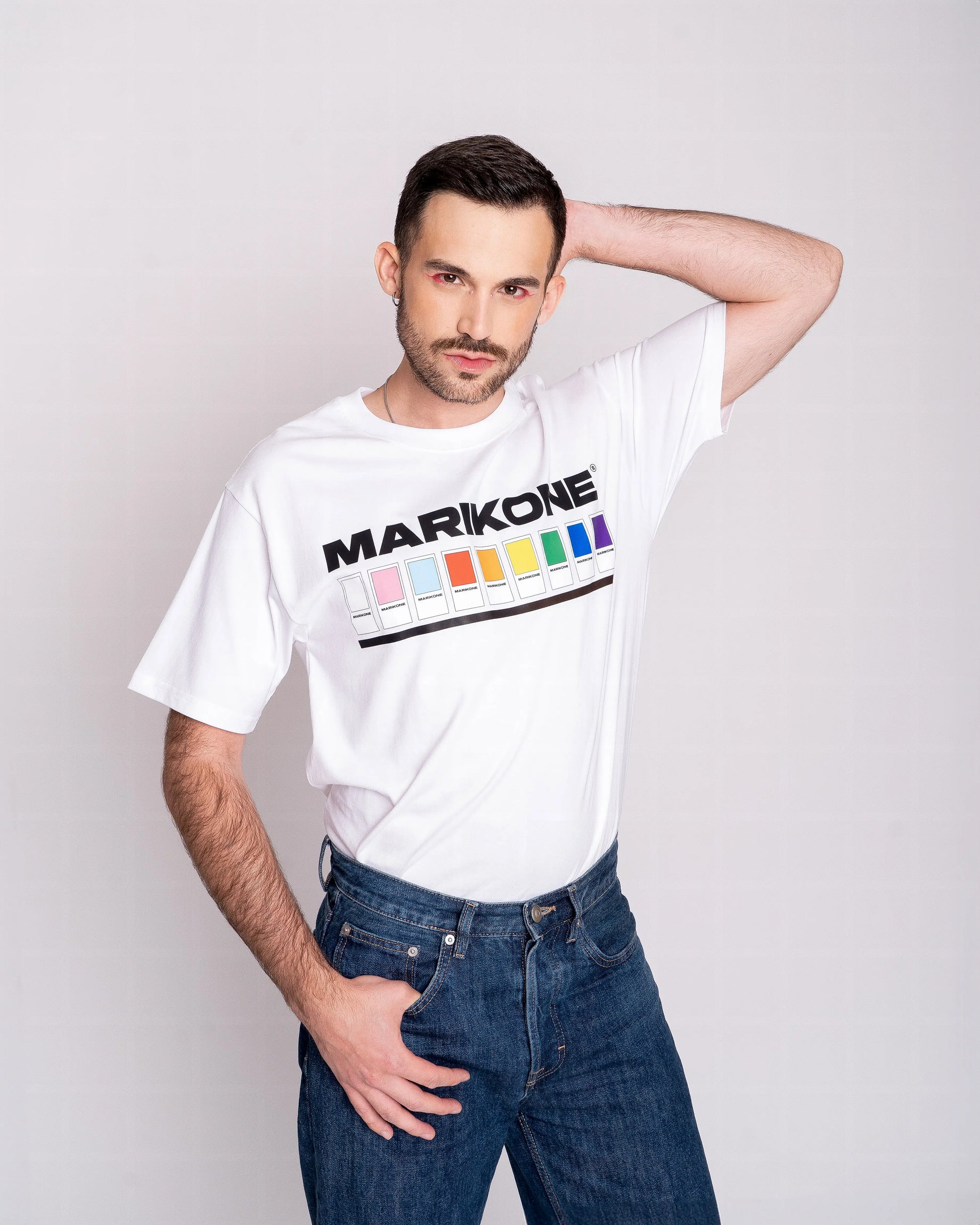 Chico con sombra de ojos rosa  posando de frente con la con la mano detrás de la cabeza y llevando una camiseta con colores de bandera gay lgtb, trans y queer con fichas de color como pantone y que dice MARIKONE