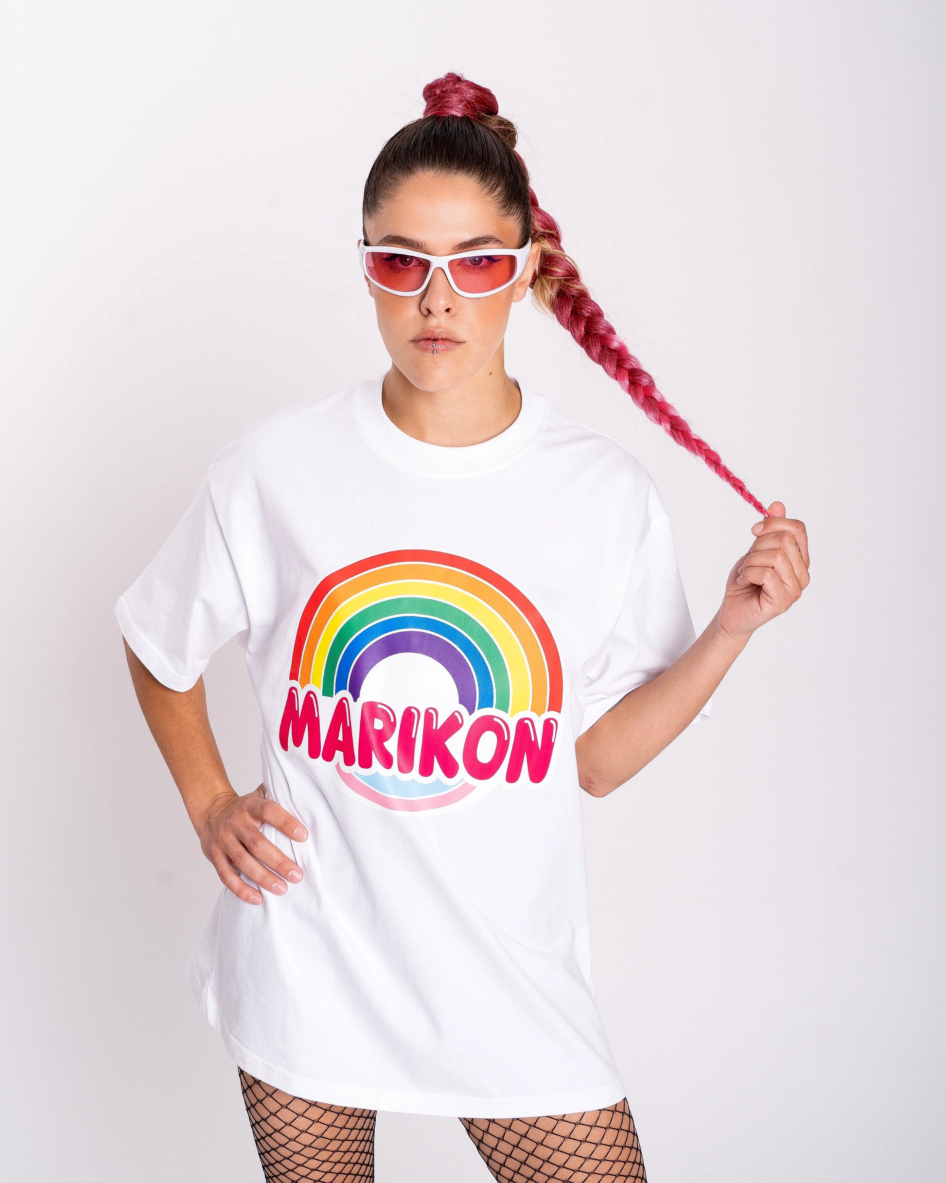 Una chica con coleta rosa que la coge por la punta. LLeva camiseta blanca con motivo MARIKON, con arcoíris y colores de bandera trans. Camiseta del orgullo y Queer.