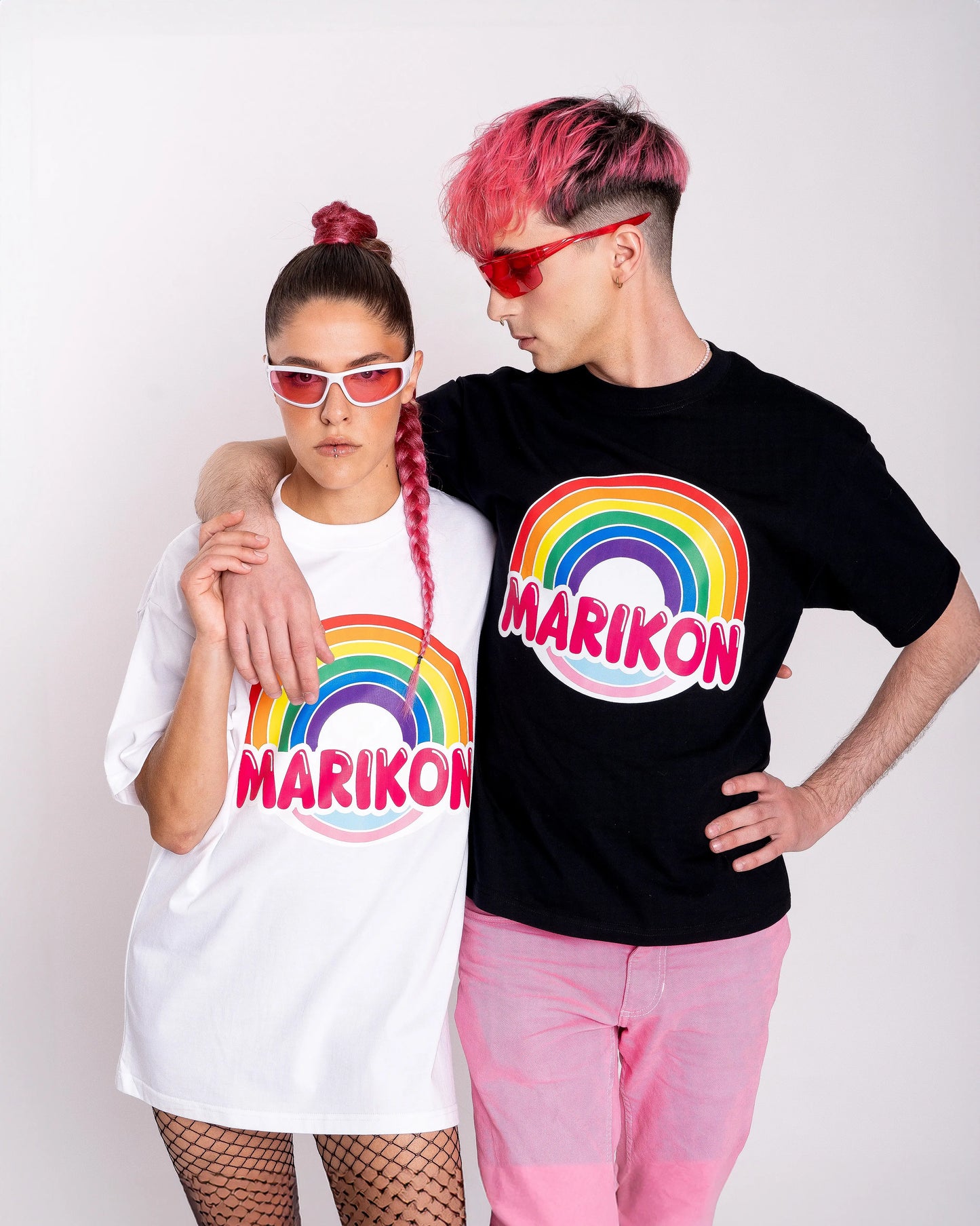 Un chico con cabello rosa abrazando a una chica con coleta rosa. Ambos llevan camisetas con motivo MARIKON, blanca y negra, con arcoíris y colores de bandera trans. Camiseta del orgullo Queer y LGTBIQ+