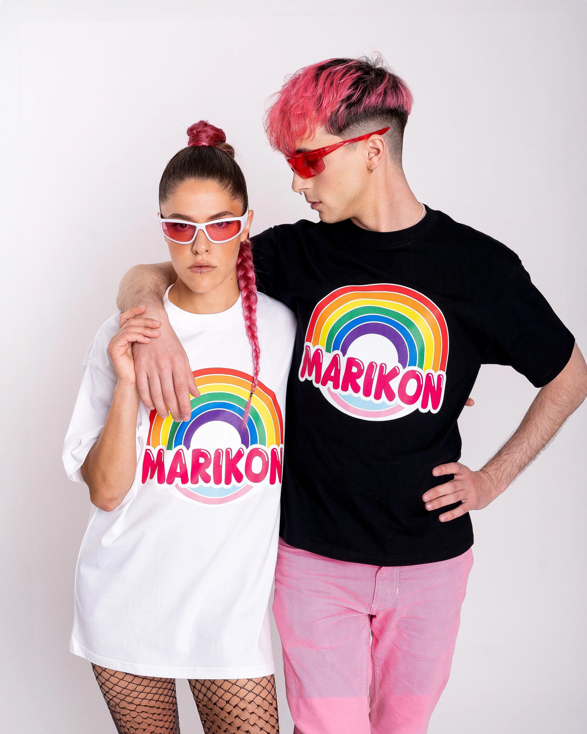 Un chico con cabello rosa abrazando a una chica con coleta rosa. Ambos llevan camisetas con motivo MARIKON, blanca y negra, con arcoíris y colores de bandera trans. Camiseta del orgullo Queer y LGTBIQ+