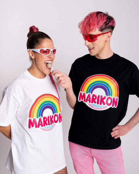 Un chico con cabello rosa dándole una piruleta a una chica con coleta rosa que la muerde con una sonrisa. Ambos llevan camisetas con motivo MARIKON, blanca y negra, con arcoíris y colores de bandera trans. Camiseta del orgullo Queer