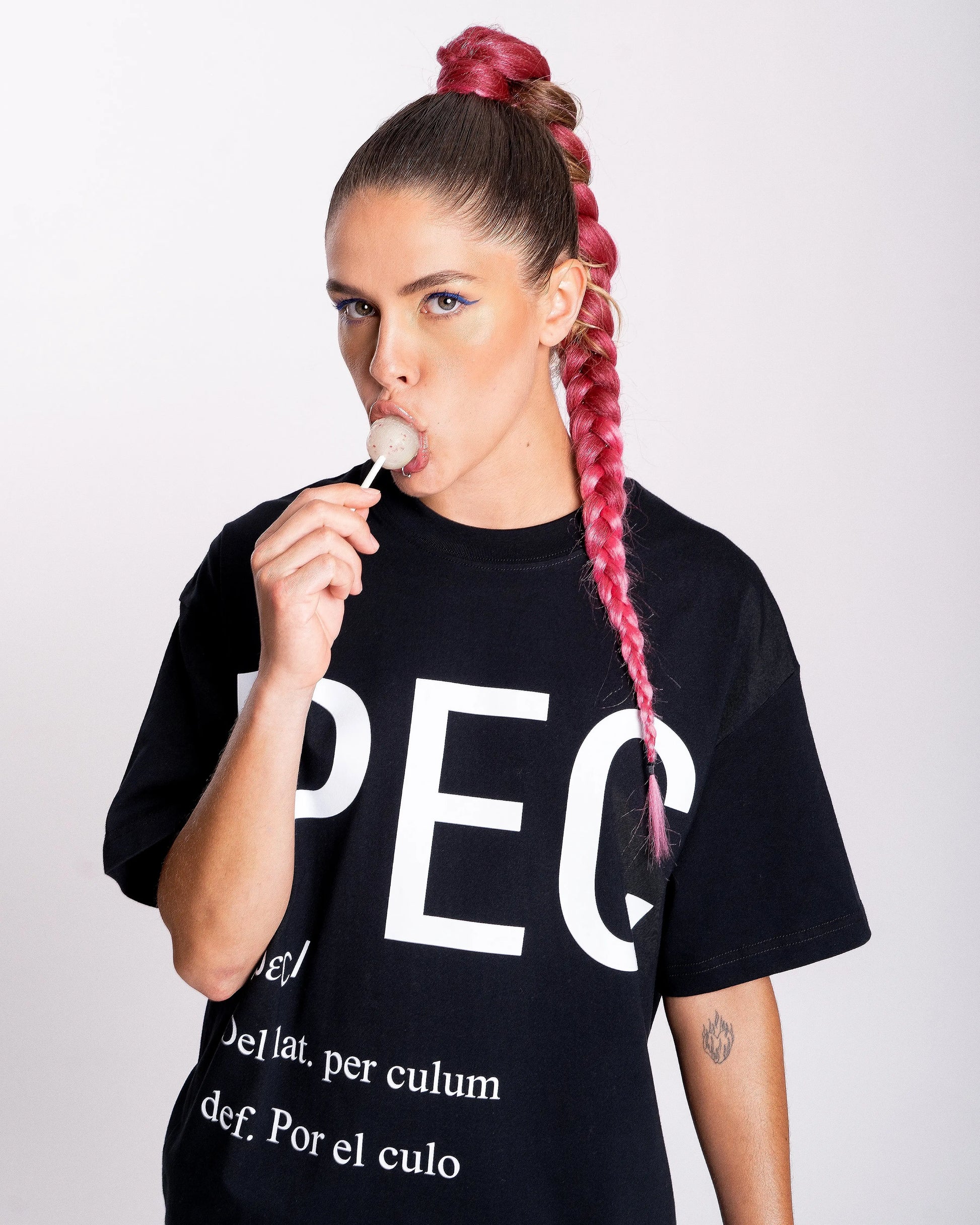 Chica queer comiendo una piruleta y lleva camiseta negra con letras blancas grandes que dicen PEC, definición: por el culo. Una expresión del colectivo LGTBIQ+, gay y Queer